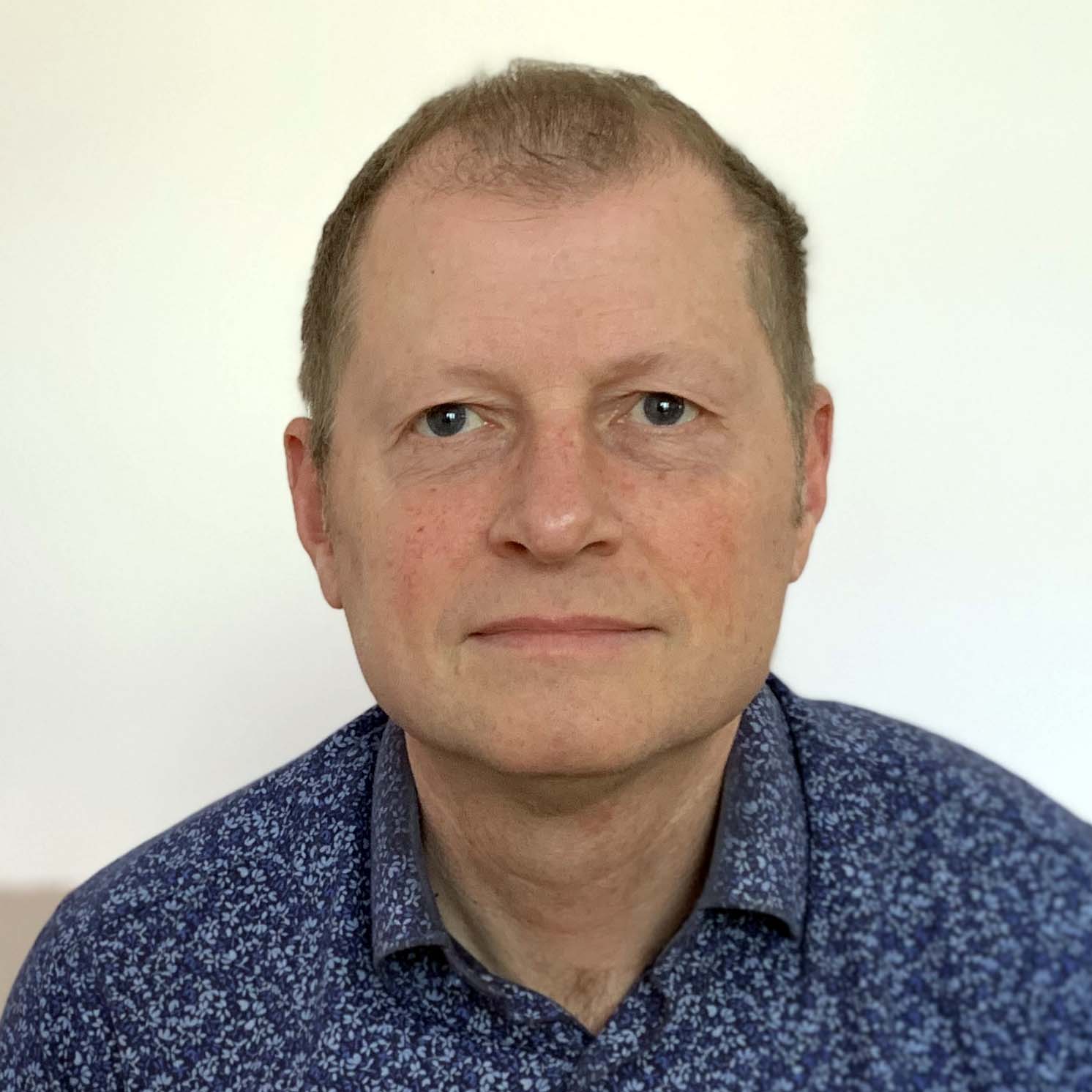 Sergio is a white man with light blonde hair and is smiling to the camera wearing a blue patterned shirt in front of a white background.